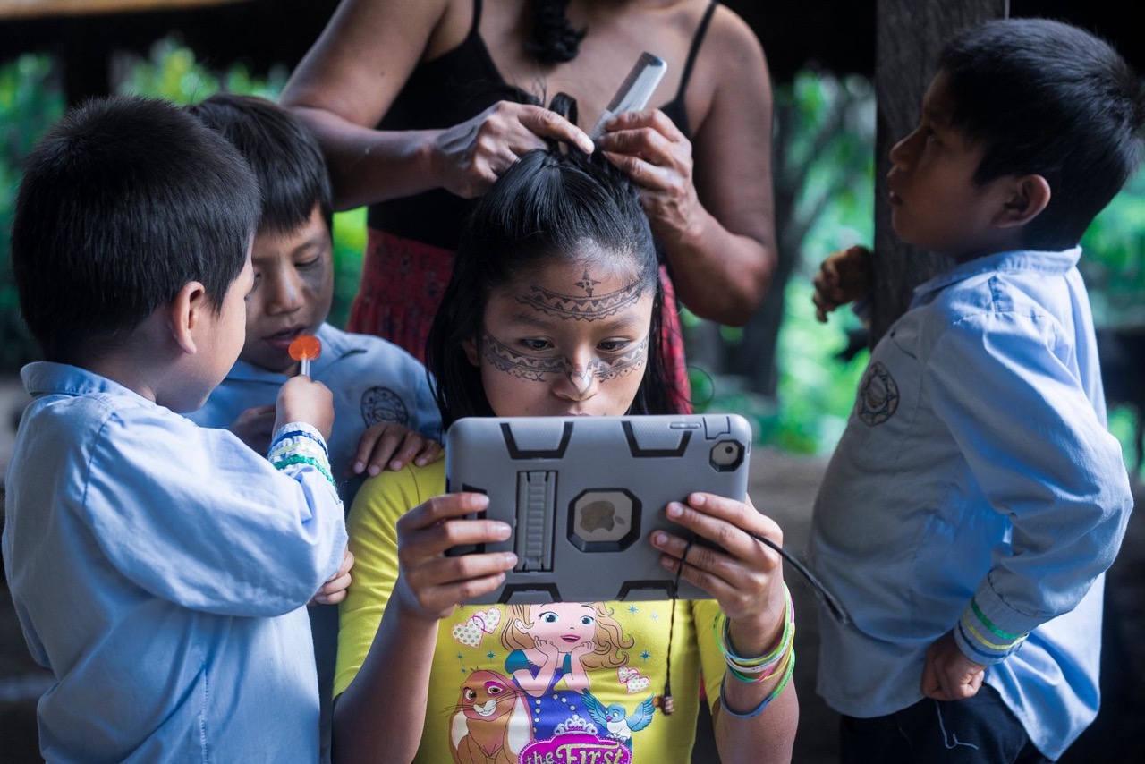 Leyli Gualinga joue sur un iPad pendant que sa mère Marcia Gualinga lui coiffe les cheveux et que son frère la regarde. - ©Misha Vallejo